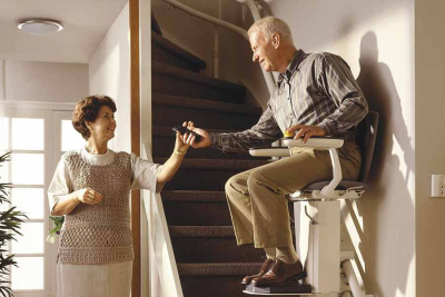 Afbeelding van een man op een witte traplift halverwege de trap en een vrouw rijkt hem de afstandsbediening van de traplift aan