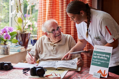 Bejaarde man die aan in zijn eigen huis aan tafel zit met potlood in hand en opengslagen schrift die begeleiding en uitleg krijgt door een thuiszorgmedewerker en een display bord van liftoffertes op tafel