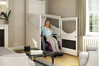 Afbeelding van een vrouw in een witte rolstoellift die uitkomt in de huiskamer die zit in een rolstoel en de liftdeur is open